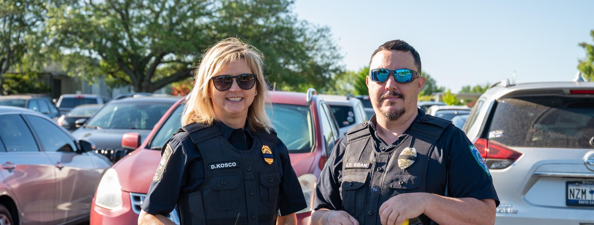 Two police officers, a woman and a man, stand side-by-side in a parking lot. They are both wearing dark blue uniforms, tactical vests, and sunglasses. The woman has blonde hair and a nameplate that reads 
