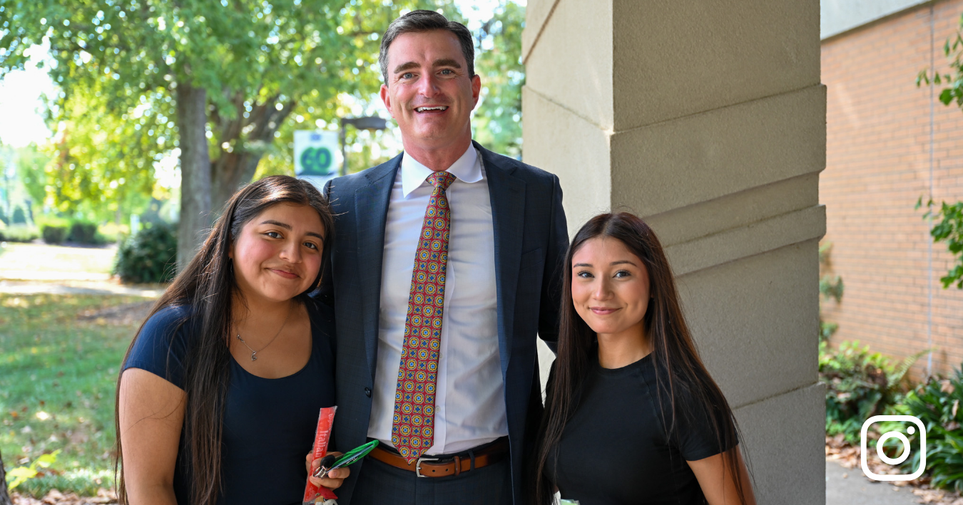 A medium shot shows a tall man in a professional dark suit and a patterned orange and blue tie smiling between two young women. They are standing outdoors near a stone pillar with lush green trees and a building in the soft-focused background.
