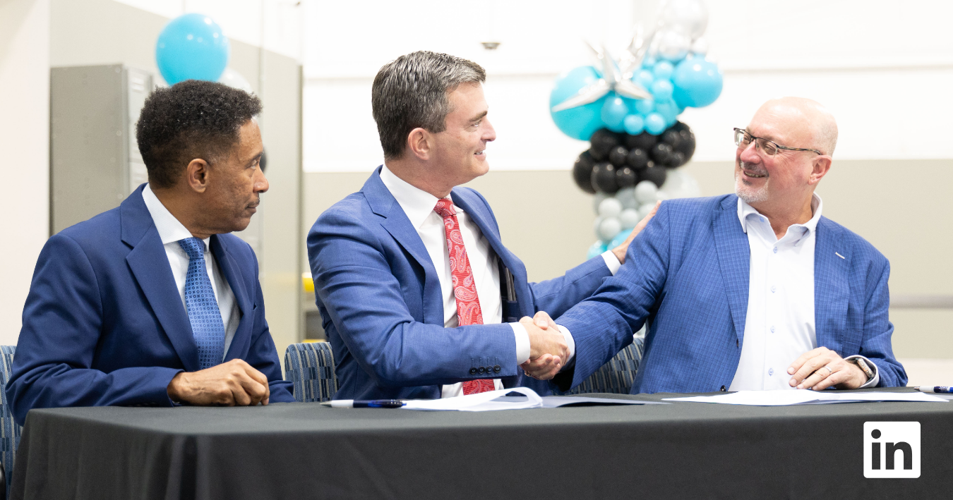 Three professionally dressed men sit at a table during a formal event, with two of them shaking hands and smiling while documents are laid out in front of them. Decorative balloons are visible in the background, suggesting a celebratory or signing occasion.