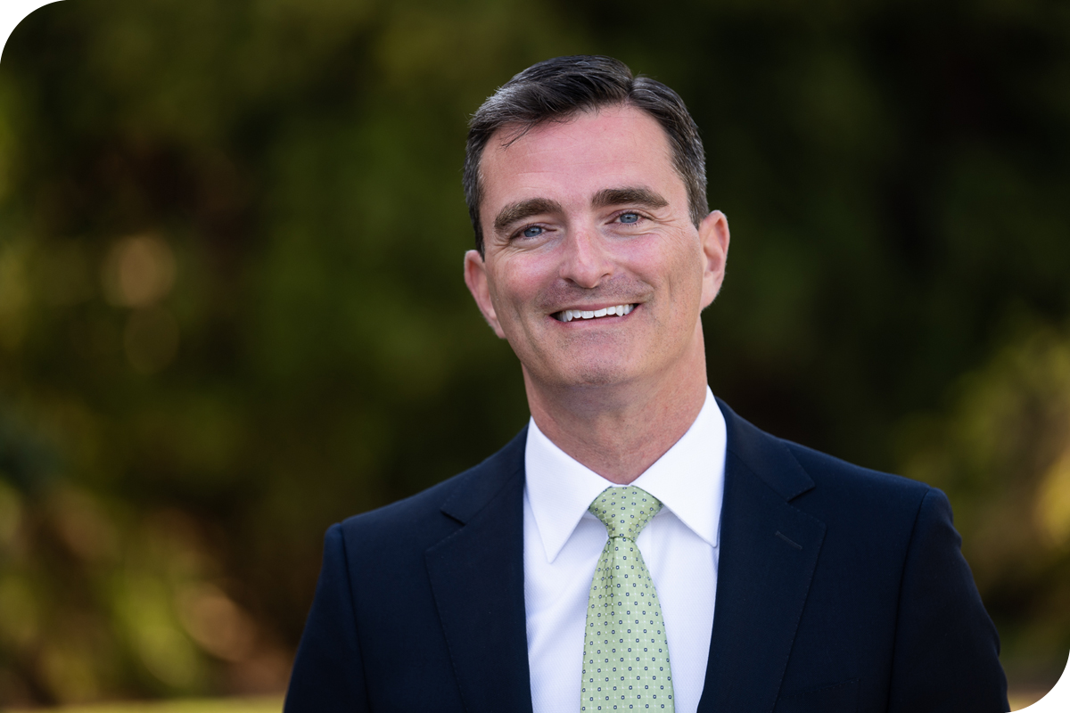 Professional outdoor headshot of an individual wearing a dark suit jacket, white dress shirt, and light green patterned tie, standing in front of a softly blurred background of green foliage, with even natural lighting highlighting facial features in a clean and polished executive-style portrait composition.