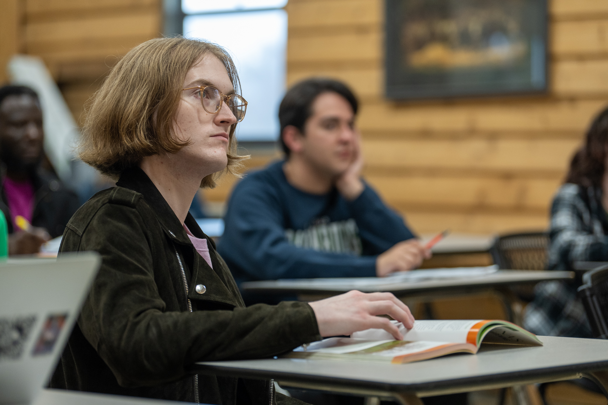 Student seated at a classroom desk follows along in an open textbook during a lecture, with additional students visible in the background. The room features wood-paneled walls, desks, and notebooks, creating an active college learning environment.