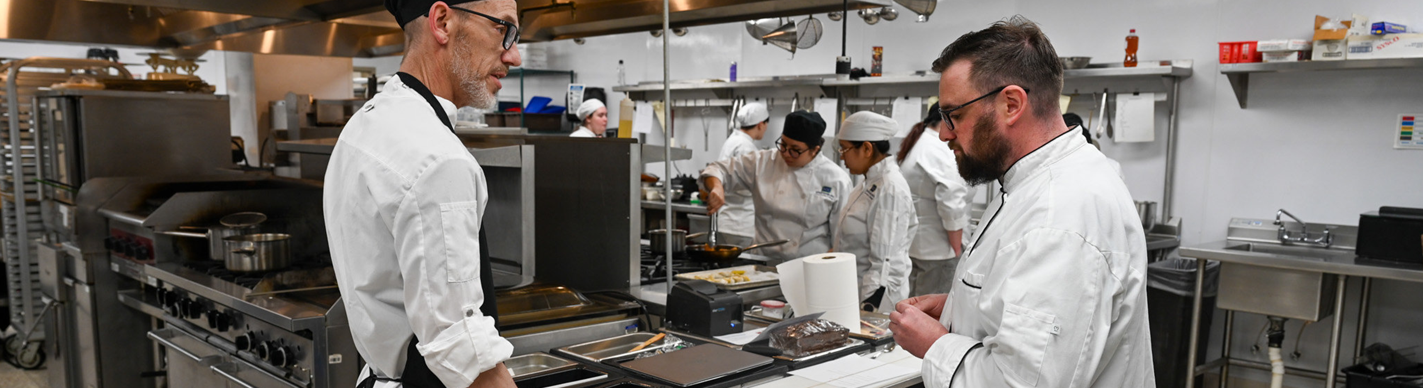 Two chefs in a commercial kitchen preparing food while several culinary students work together in the background, surrounded by professional cooking equipment and stainless-steel surfaces.