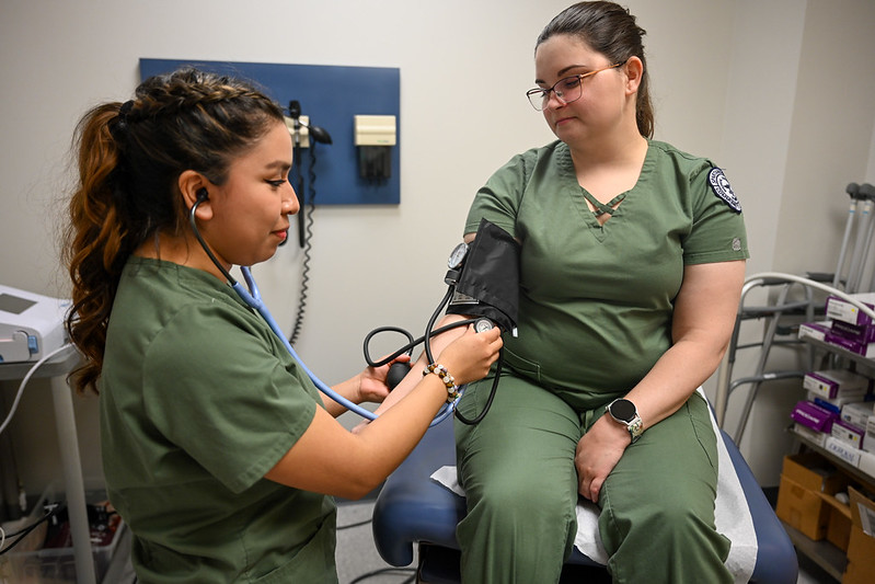 A medical assisting student checks another student’s blood pressure using a stethoscope and cuff in a clinical lab setting while both wear green medical scrubs