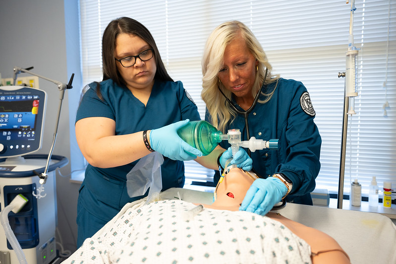 Two healthcare students in dark teal scrubs operate a bag valve mask resuscitator and use a stethoscope on a medical simulation mannequin in a training room.