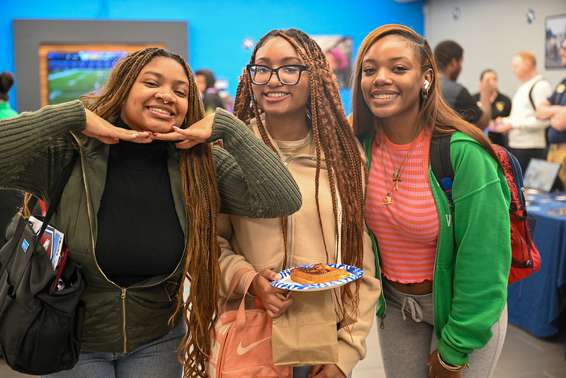 Three smiling students pose together at a campus event inside a student center. One student playfully rests her chin on her hands, while another holds a paper plate with a pastry. The students wear casual clothing and backpacks, with tables, displays, and other attendees visible in the background.