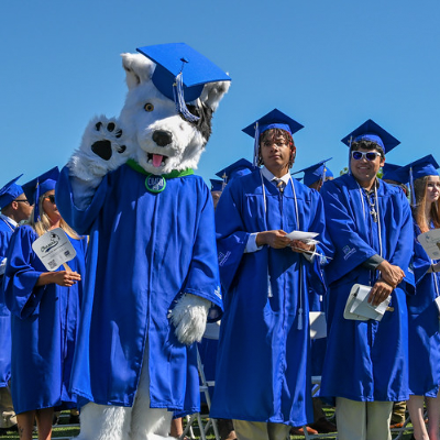 The image shows Chaser, the official Spartanburg Community College mascot, dressed in a royal blue graduation gown and cap, waving alongside a group of SCC graduates at a commencement ceremony.