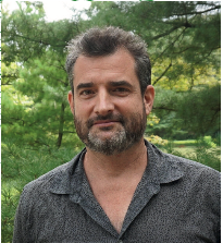 Portrait of a bearded man with short dark hair wearing a gray patterned short-sleeve button-up shirt, standing outdoors in front of green trees and foliage, looking at the camera with a slight smile.