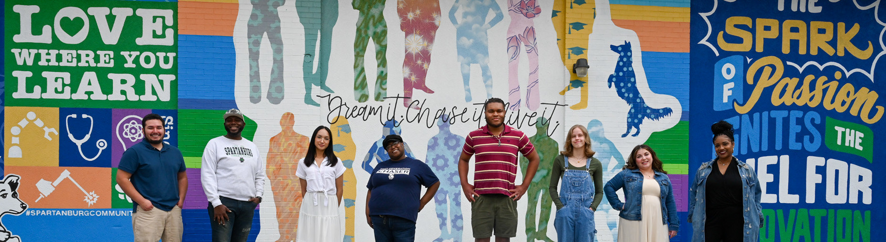 Group of students stands in a line in front of a large colorful mural at Spartanburg Community College featuring bold graphics, silhouettes, and text including “LOVE WHERE YOU LEARN,” “Dream it. Chase it. Live it,” and “The spark of passion ignites the fuel for innovation.” The students pose on a paved walkway with the vibrant campus artwork filling the background.