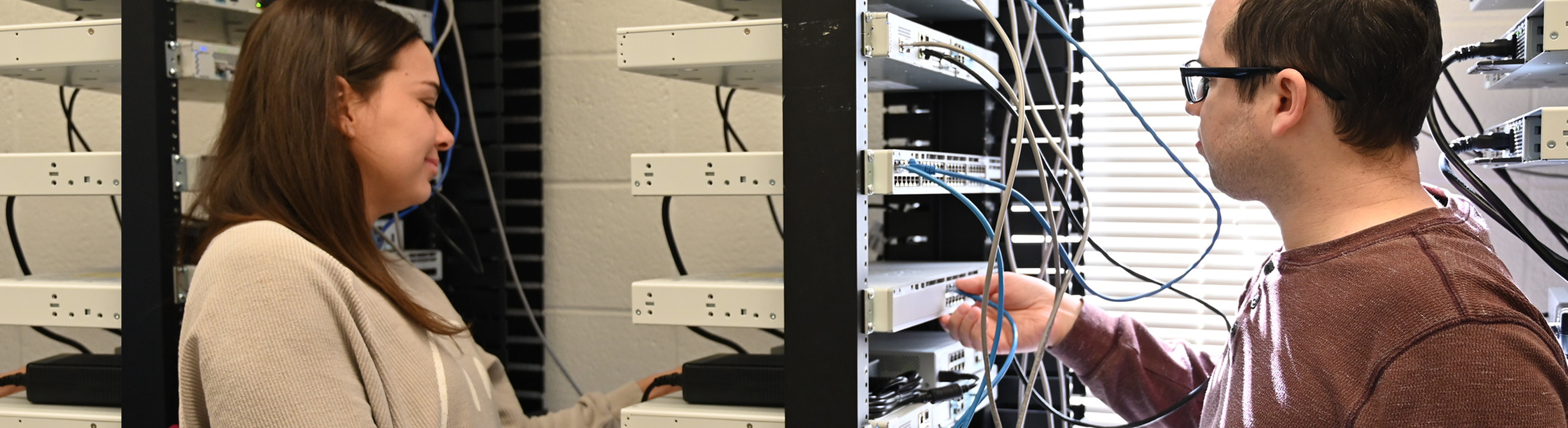Two individuals work at an equipment rack in a networking or cybersecurity lab, connecting cables and configuring network hardware. One inserts a blue Ethernet cable into a switch while the other stands nearby, with multiple network devices, patch panels, and cables arranged in the rack, indicating hands-on IT infrastructure training.