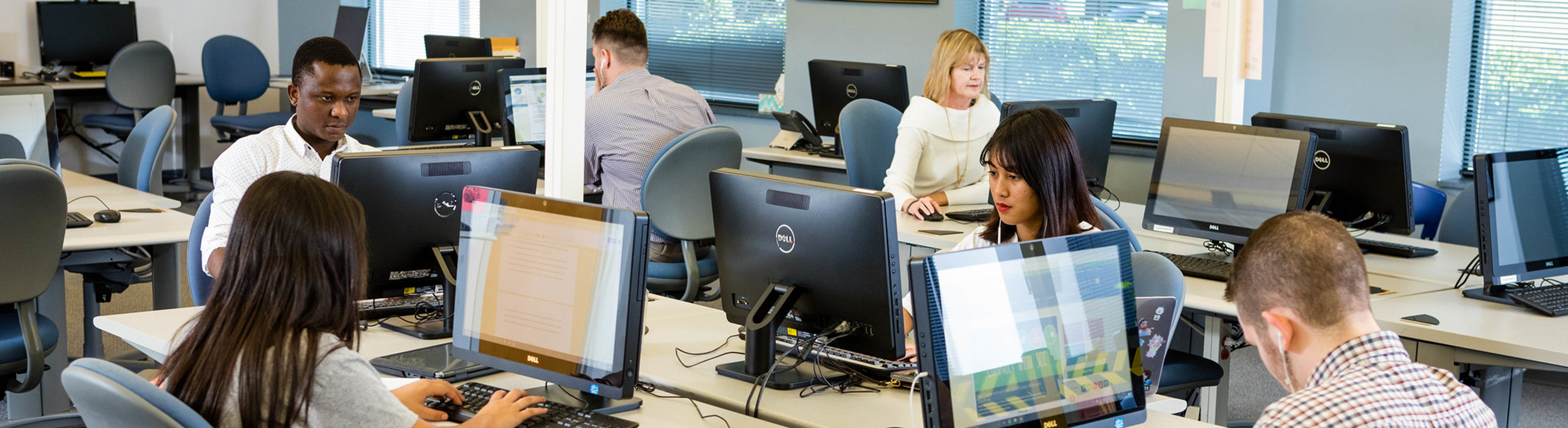 Students seated at desktop computers in a classroom lab work individually on assignments while facing monitors arranged in rows. The room includes multiple workstations, rolling chairs, large windows with blinds letting in natural light, and an instructor or additional participants visible in the background, indicating a business or computer-based learning environment.