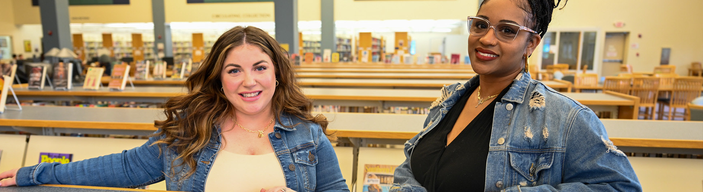 Two individuals stand together inside a college library, leaning against a railing with bookshelves and rows of seating visible behind them. Both wear denim jackets and face the camera, with a bright, open study space and organized book displays creating a welcoming academic environment.