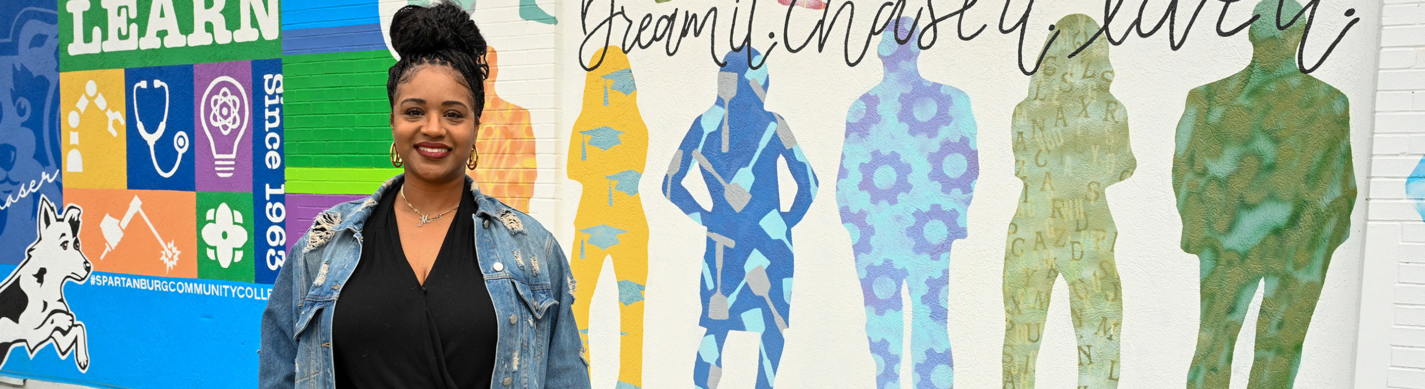 Individual stands outdoors in front of a colorful mural at Spartanburg Community College featuring silhouettes and bold text including “Learn” and “Dream it. Chase it. Live it.” The mural includes icons related to science, healthcare, and education, with SCC branding elements visible on the left side.