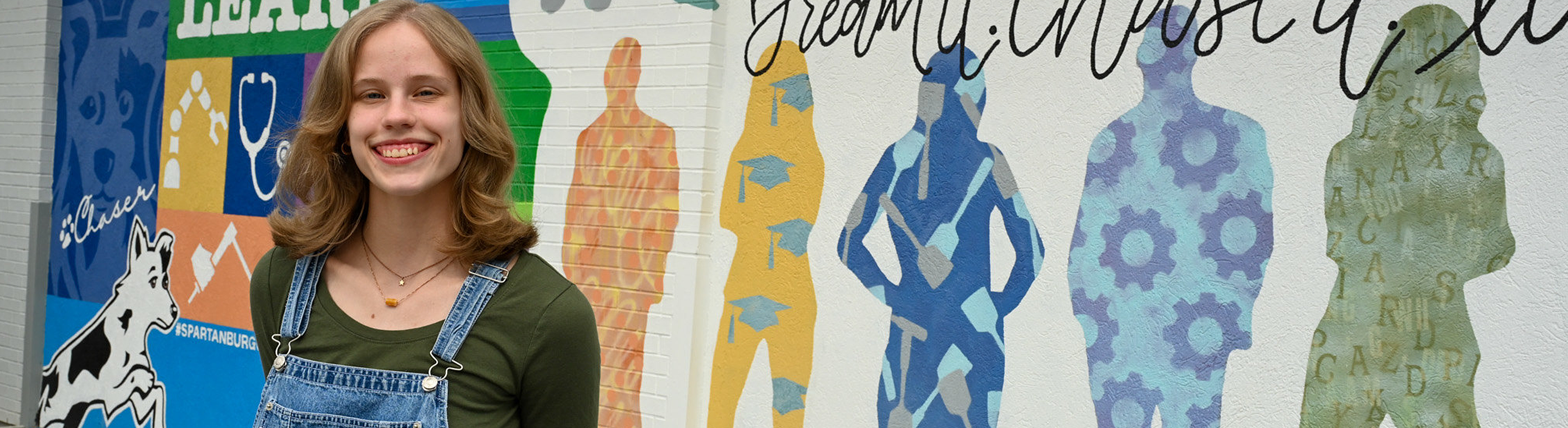 Individual standing outdoors in front of a colorful Spartanburg Community College mural featuring silhouettes and bold text including “Dream it. Chase it. Live it.” and “Learn.” The mural displays icons representing education and career fields, while the individual faces the camera against the vibrant campus backdrop.