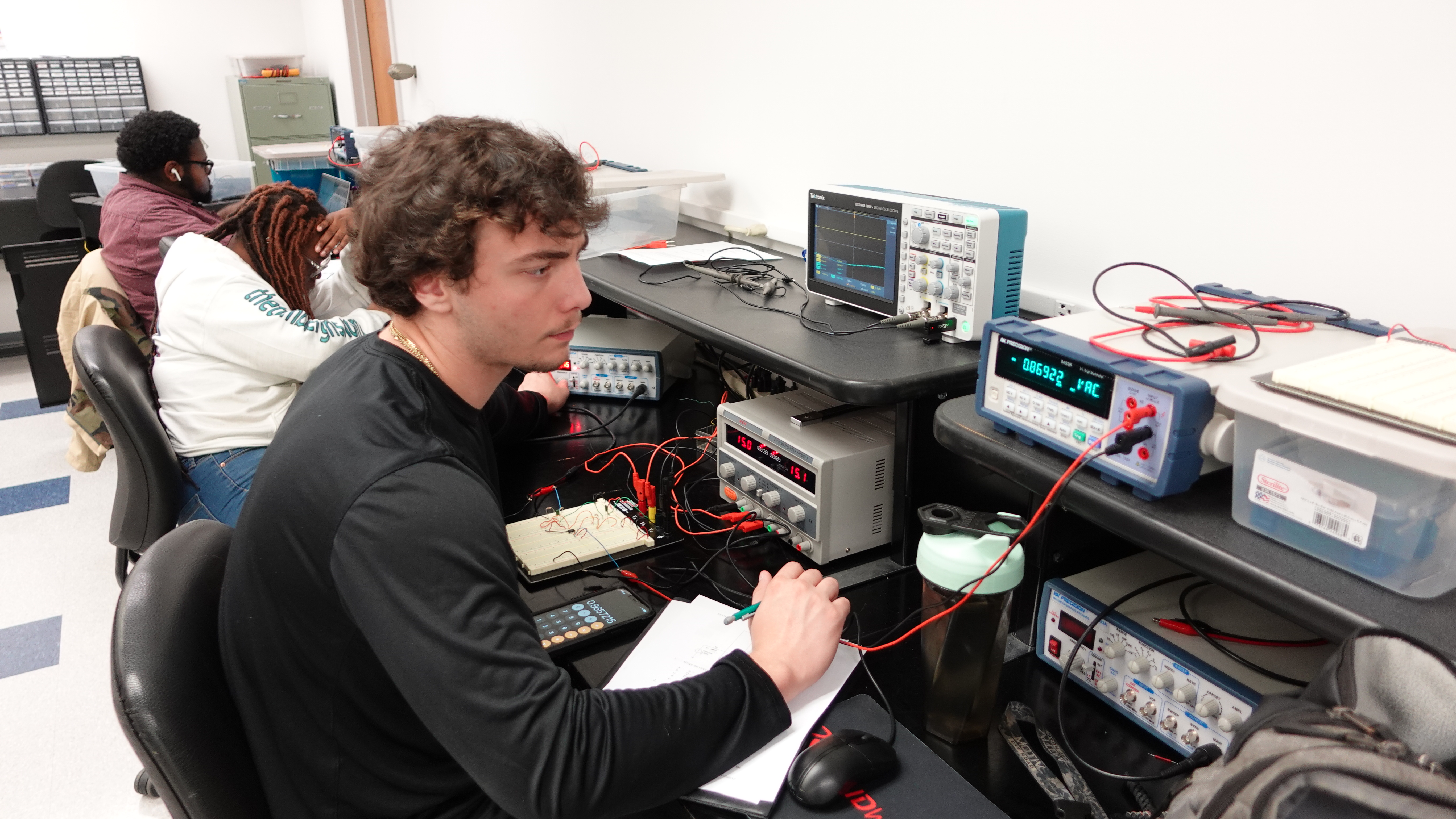 Students seated at a workstation in an electronics lab work with circuit boards and testing equipment, including an oscilloscope and power supply. One writes notes while interacting with wired components on a breadboard, while others work at nearby stations, indicating a hands-on electrical or engineering training environment.