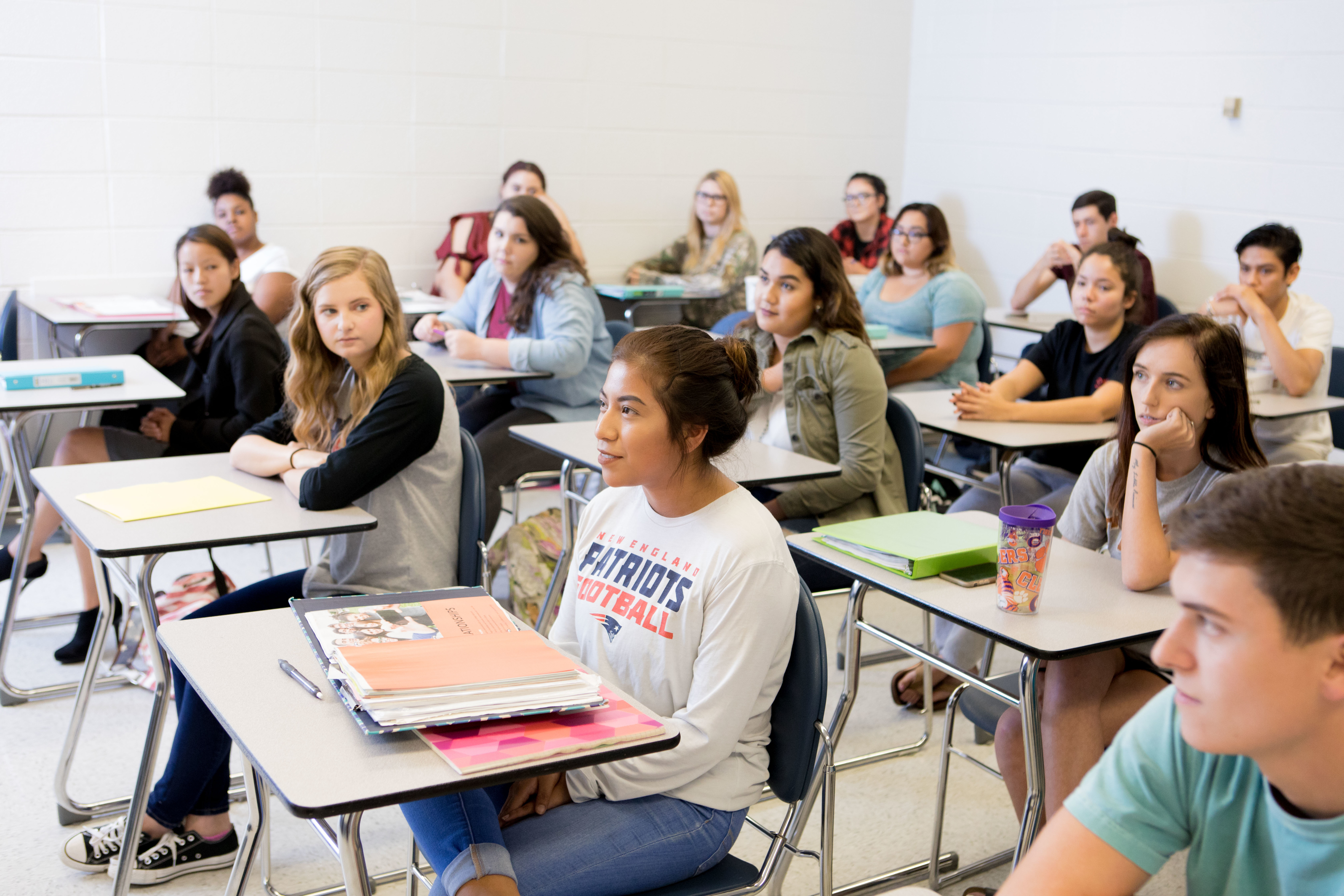 Students seated at individual desks in a classroom listen during a lecture, with notebooks, binders, and personal items visible on desks throughout the room. The classroom features rows of desks, bright lighting, and a mix of students facing forward in an active college learning environment.