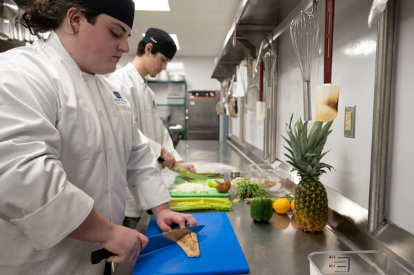 Culinary students in chef uniforms work at stainless steel preparation stations in a commercial teaching kitchen, chopping vegetables and preparing ingredients on cutting boards. Fresh produce, including celery, peppers, citrus fruits, and a pineapple, is arranged on the counter, with kitchen equipment and utensils visible throughout the training environment.