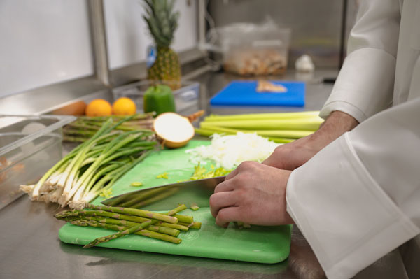 Close-up view of a culinary student chopping vegetables on a green cutting board in a commercial teaching kitchen. Fresh ingredients including asparagus, green onions, celery, peppers, citrus fruits, and a pineapple are arranged on a stainless steel prep station, with additional cutting boards and food preparation materials visible in the background.