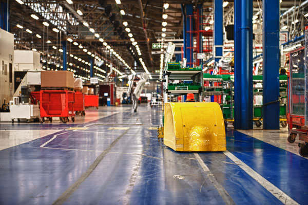 Interior of a large manufacturing facility with a wide production aisle running through the center, surrounded by industrial equipment, storage racks, carts, and workstations. Bright overhead lighting, blue support columns, and organized assembly areas create a modern logistics and manufacturing environment.