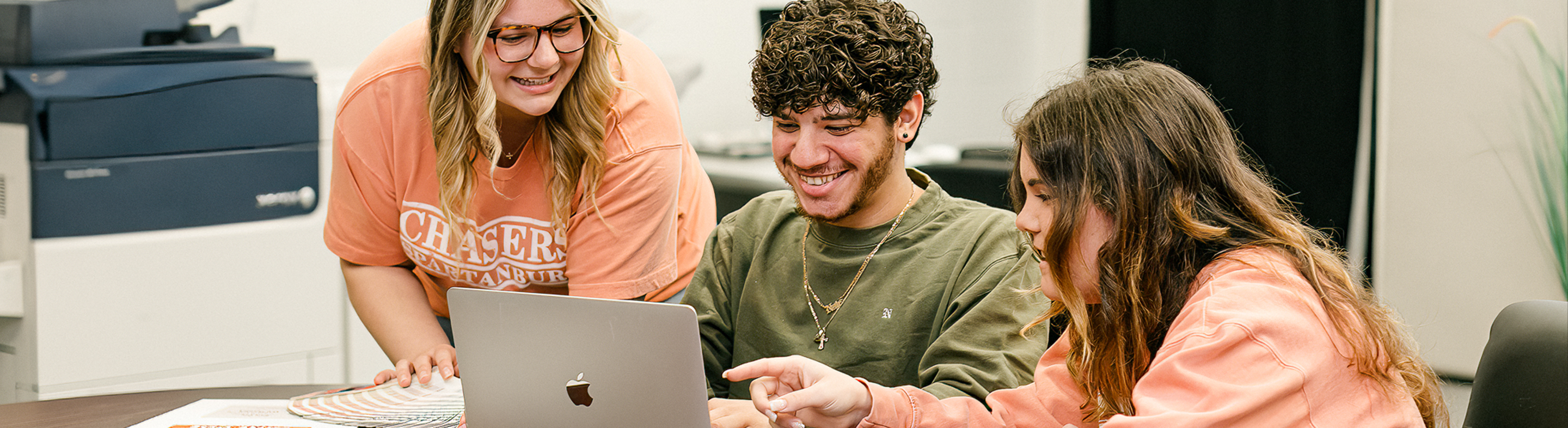 Three students collaborate around a laptop, smiling and pointing at the screen while working together in a classroom setting.