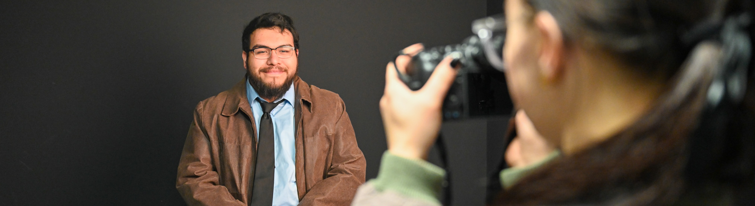 A photographer takes a portrait of a smiling man wearing glasses, a blue shirt, tie, and brown jacket, seated against a dark background.