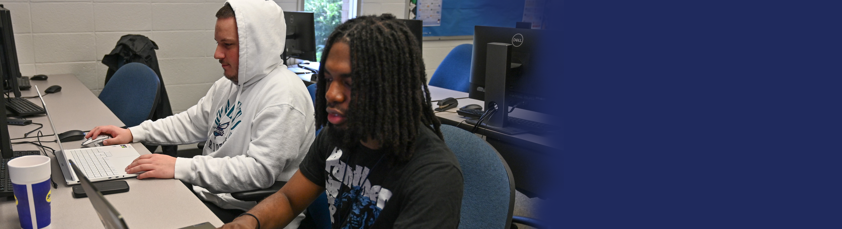 Two young men are sitting at a long desk in a computer lab or classroom. One man on the left wears a white hoodie and uses a silver laptop, while the man on the right, with long dreadlocks and a black t-shirt, works on another computer. Several desktop monitors and a blue-and-white cup are visible on the desk.