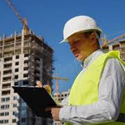 Individual wearing a white hard hat and high-visibility safety vest stands at a construction site holding a clipboard and reviewing documents. A multi-story building under construction with scaffolding and cranes is visible in the background, indicating an active construction or project management environment.