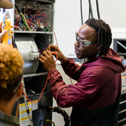 Individual works on an open electrical unit, using a tool to adjust wiring connections inside a panel filled with multicolored cables and internal components. Another person stands nearby observing, with equipment and wires exposed, indicating hands-on electrical repair or technical training in a workshop environment.