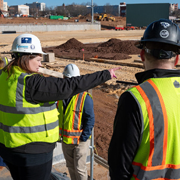 Group of individuals wearing hard hats and high-visibility safety vests stand together at a construction site, with one gesturing while discussing an area under development. The setting includes graded dirt, construction materials, and work zones, indicating a hands-on construction training or supervision environment.