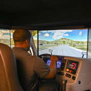 Individual seated in a driving simulator holds a steering wheel while facing a large curved screen displaying a roadway scene with hills, guardrails, and a multi-lane path. The console includes buttons, gauges, and a digital display, indicating a commercial driver training or truck driving simulation environment.