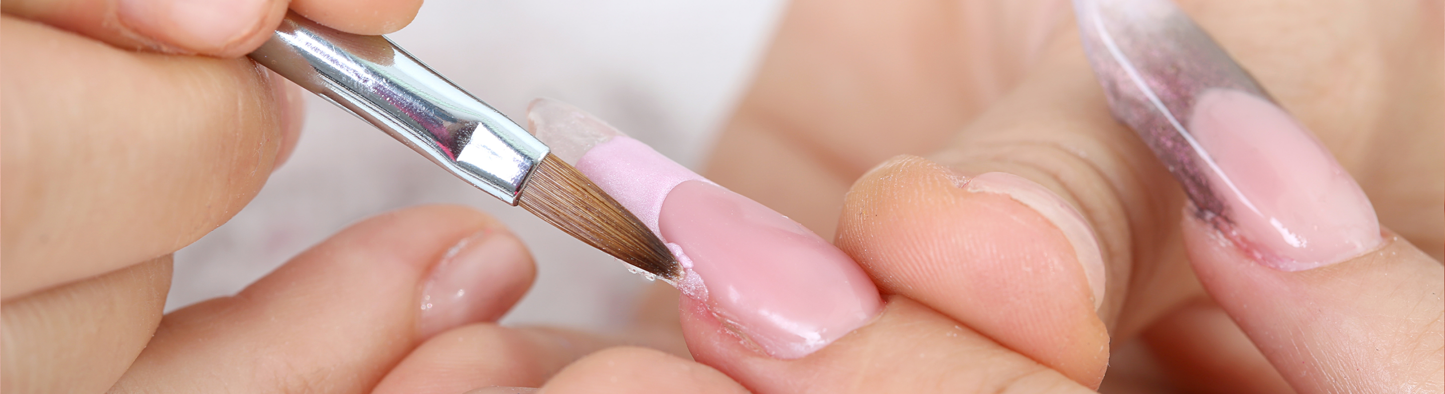 Close-up view of a cosmetology procedure showing a small brush applying pink acrylic or gel product onto a shaped fingernail while another tool refines a neighboring nail. The process takes place in a clean salon or training setting with detailed focus on nail shaping and application.