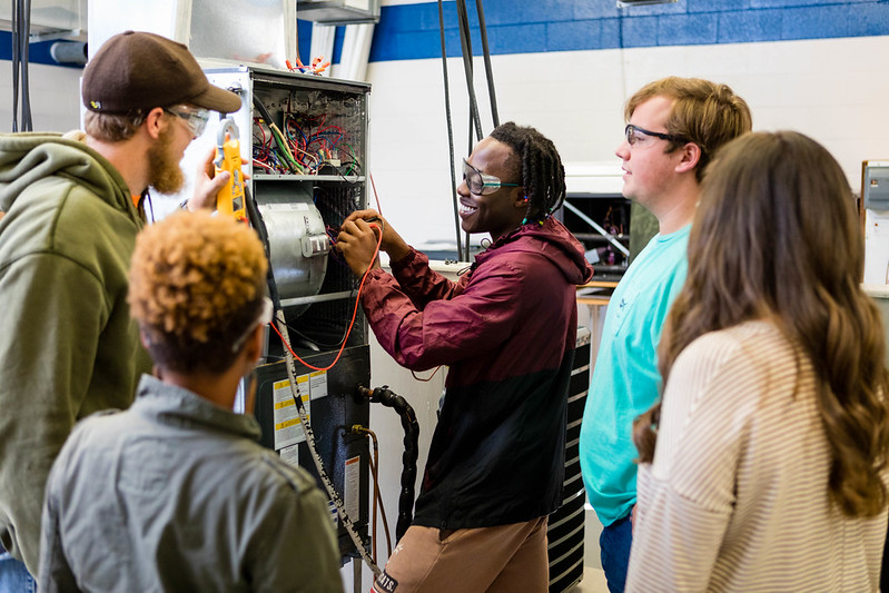 A diverse group of five students in a technical workshop focused on a piece of HVAC machinery. A student in a maroon jacket smiles while using a multimeter to test exposed wiring, while another student in a brown cap and safety glasses assists from the left. Three other students look on attentively. The setting is a brightly lit classroom with white and blue walls, emphasizing a hands-on, collabor