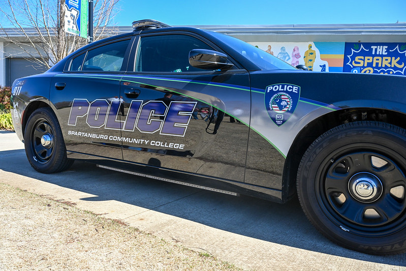 Side view of a black Spartanburg Community College police vehicle parked outdoors, featuring large white “POLICE” lettering and the SCC logo on the door. The car is positioned on a paved surface with a colorful campus mural and building visible in the background under a clear blue sky.