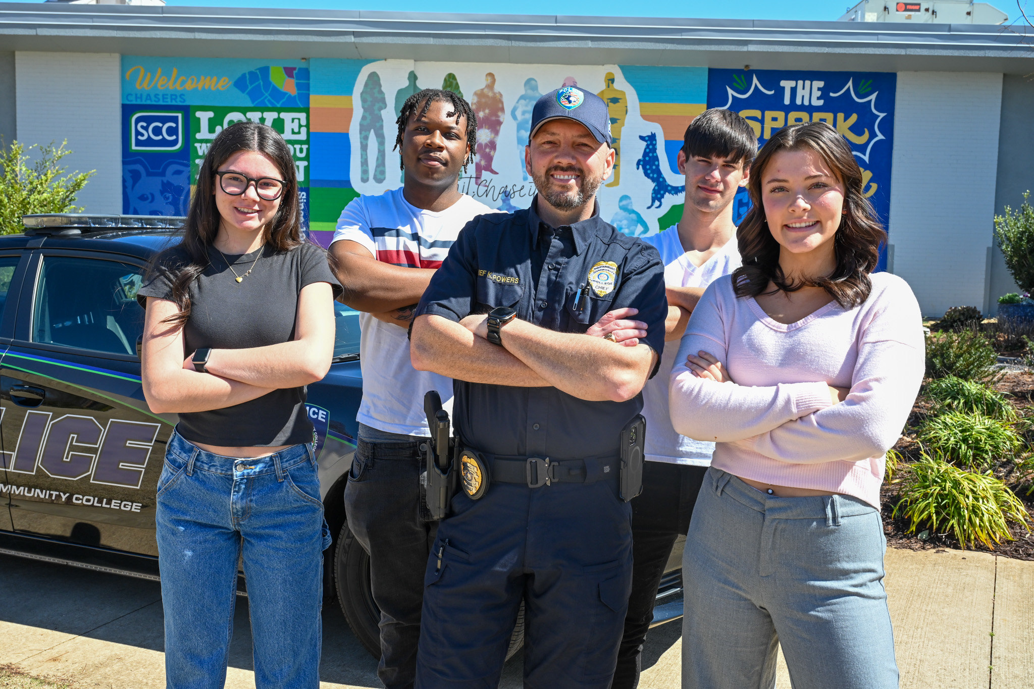 Group of five individuals standing outdoors in front of a Spartanburg Community College mural and a police vehicle, posing with arms crossed. One individual wears a law enforcement uniform with visible badge and equipment, while others wear casual clothing, with SCC branding and bright colors visible on the mural in the background.