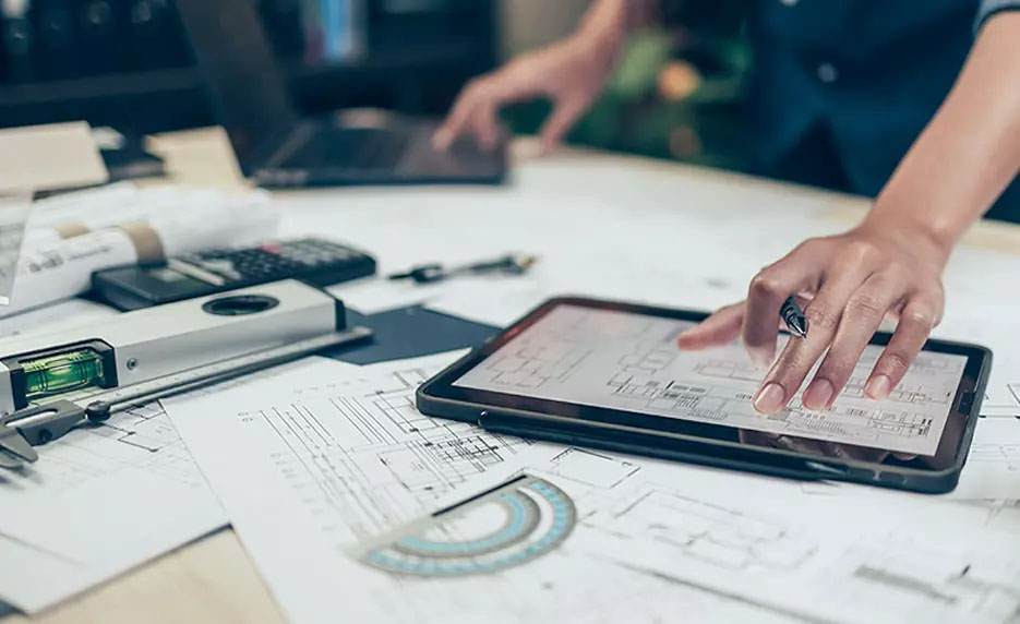 Close-up of a workspace showing a tablet displaying architectural floor plans while a hand holds a pen over the screen. Surrounding the tablet are printed blueprints, a calculator, measuring tools, and rolled plans, with another workstation and laptop blurred in the background, indicating construction planning or estimation work.