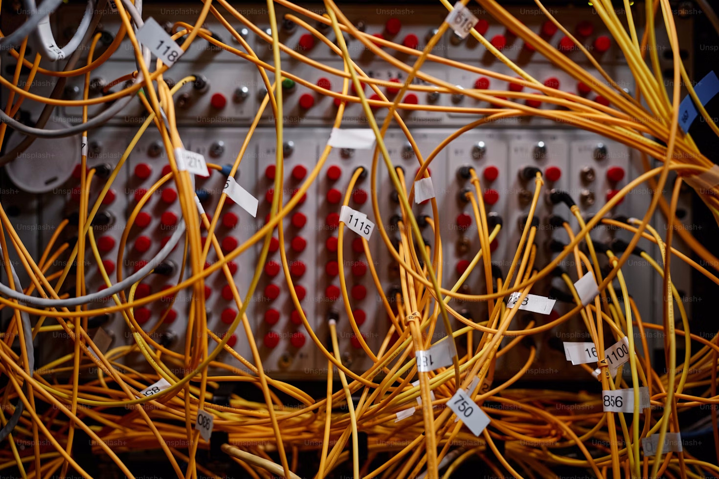 Close-up view of a dense network of yellow electrical wires connected to a control panel filled with rows of red and metallic connection points. Several wires have small white labels with numbers, and the panel features multiple switches and ports, indicating a complex electrical or industrial system.