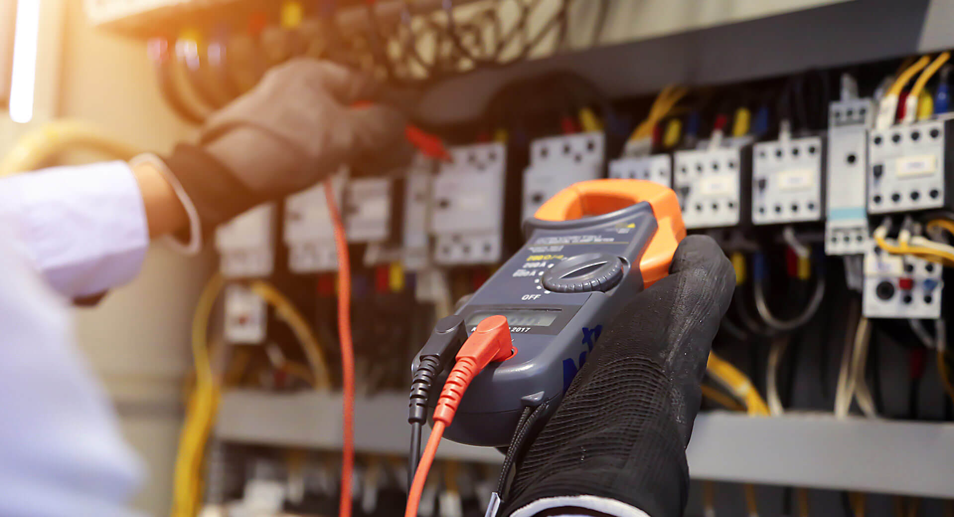 Close-up view of a technician wearing black protective gloves using a digital multimeter to test electrical components inside an open industrial control panel. The background shows multiple relays, wiring connections, and color-coded cables arranged across the panel, indicating electrical diagnostics or maintenance work.