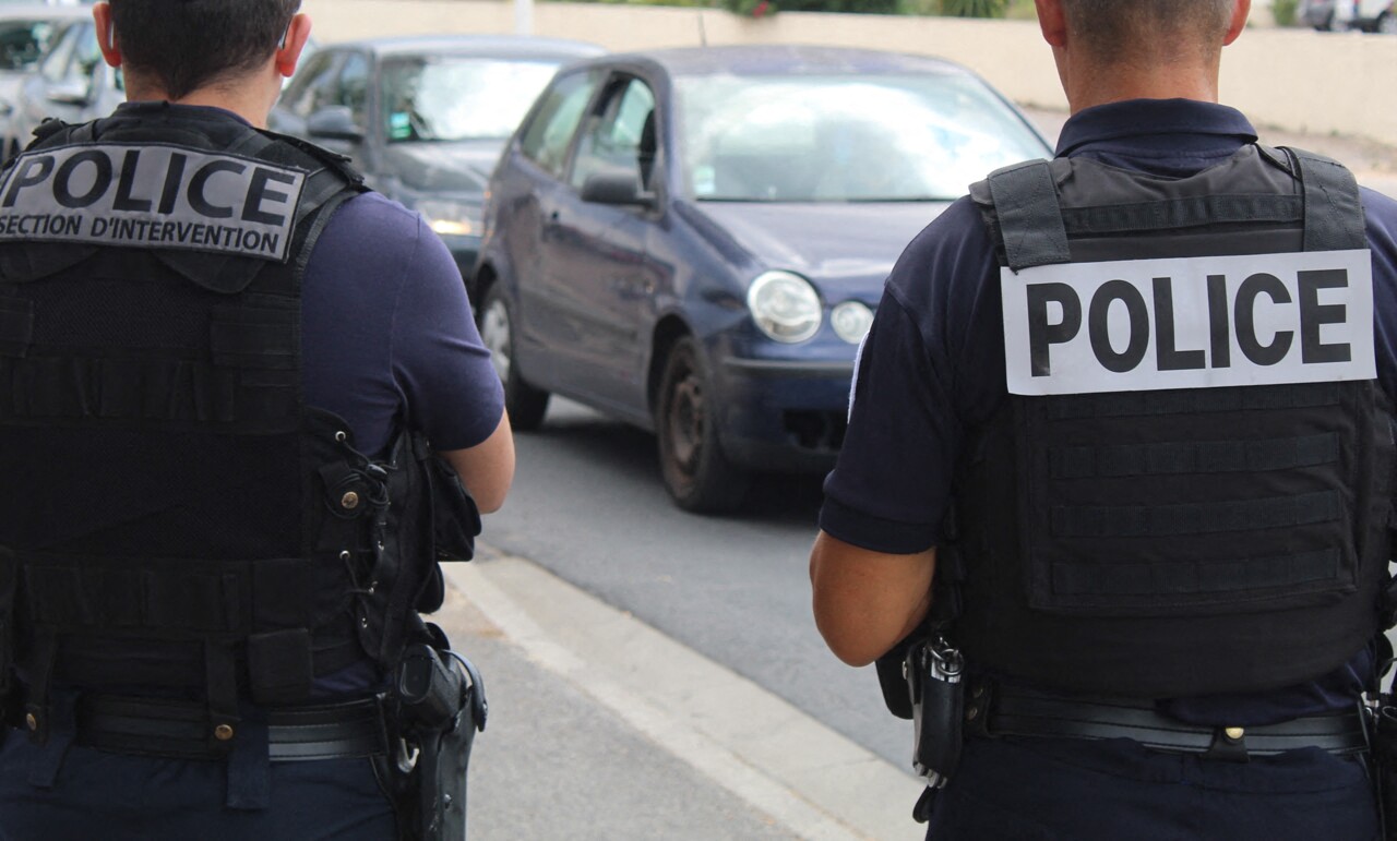 Two police officers wearing protective vests labeled “POLICE” stand on a street facing a stopped vehicle during a roadside interaction. The scene includes multiple cars on the road, with one vehicle closest to the officers, and an urban street environment visible in the background.