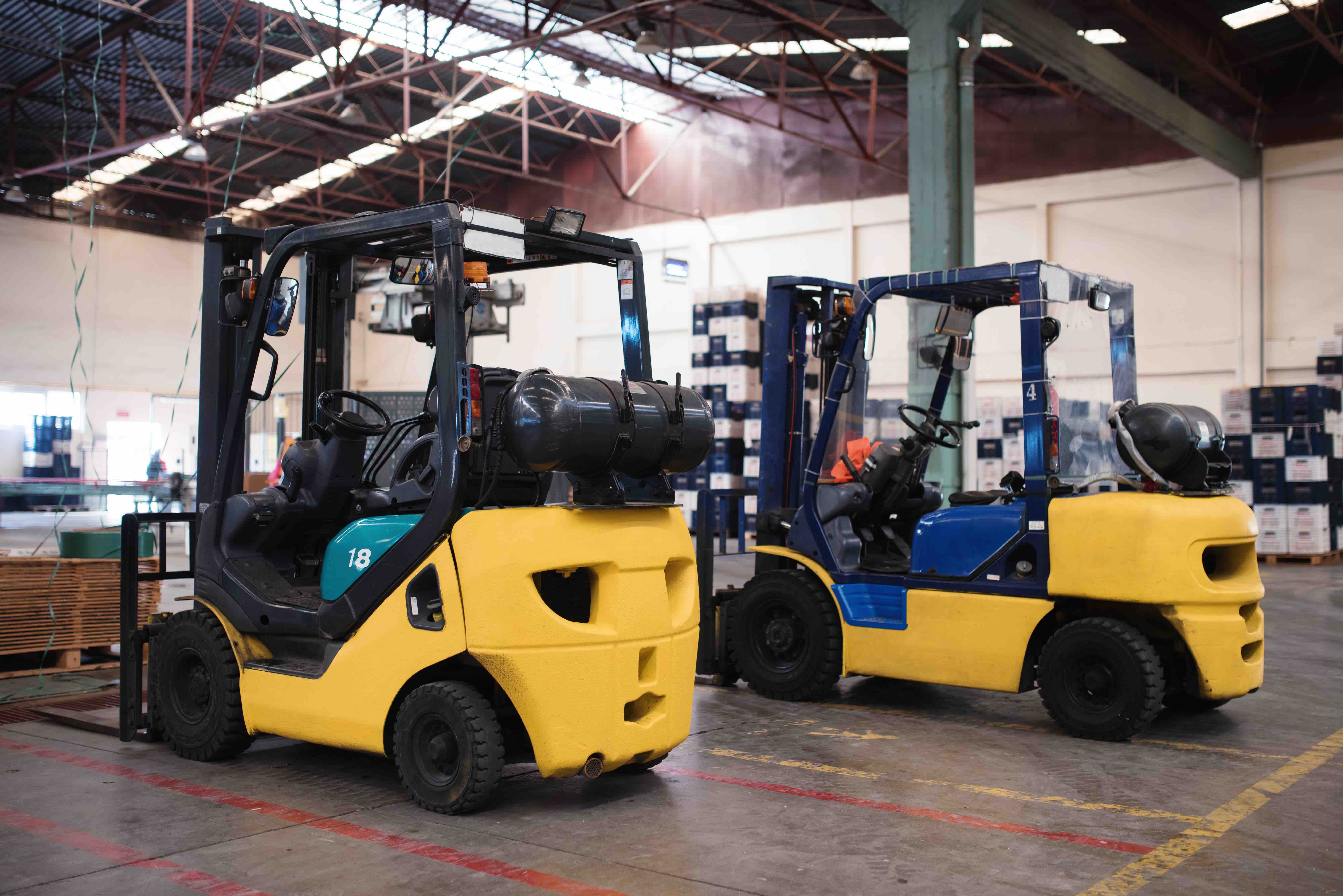 Two forklifts are parked side by side inside a large industrial warehouse with high ceilings, metal beams, and overhead lighting. The forklifts are yellow and blue, with visible propane tanks mounted on the back, and pallets and stacked boxes can be seen in the background, indicating a material handling or logistics environment.