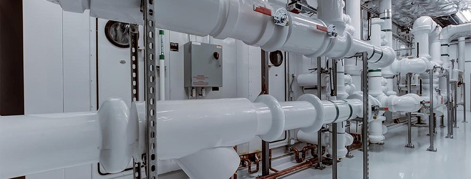 A mechanical room filled with complex industrial HVAC piping systems. Large, insulated white pipes run horizontally and vertically across the room. They are supported by metal struts and brackets. An electrical control box sits on the wall behind the pipes. The entire room has a sterile white floor and ceiling.