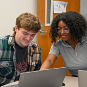 Two students collaborating on laptops in a computer lab classroom while a humanoid robot sits nearby, highlighting teamwork, technology, and STEM education