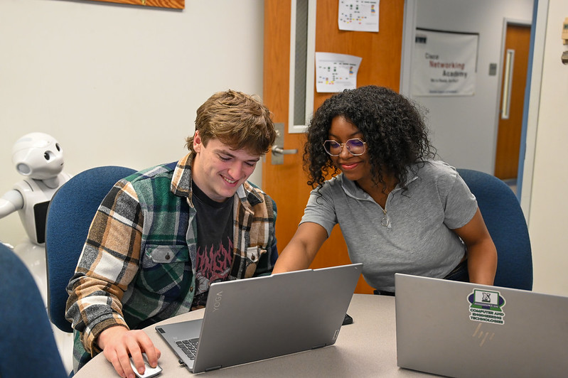 Two students collaborating on laptops in a computer lab classroom while a humanoid robot sits nearby, highlighting teamwork, technology, and STEM education
