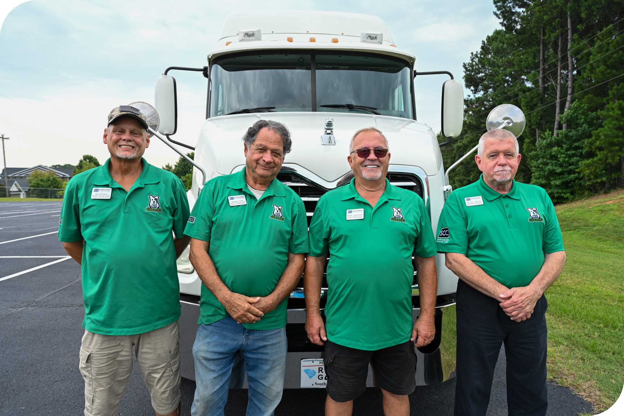 Four men stand confidently in a row, all wearing matching bright green polo shirts featuring an embroidered logo. They are positioned in front of the large, white grille of a Mack truck, which serves as a clean, industrial backdrop. The setting appears to be an outdoor asphalt parking area under a bright, overcast sky, with lush green trees visible in the distance to the right.