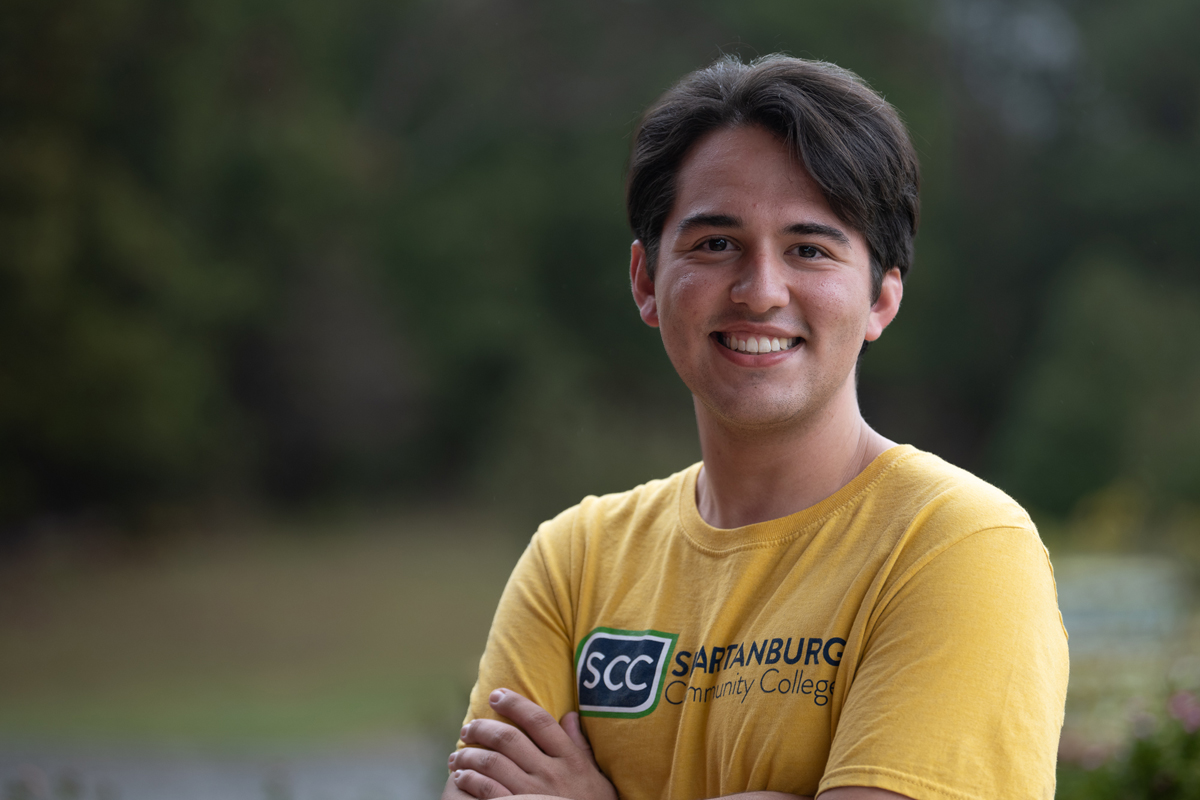 Outdoor portrait of an individual wearing a yellow Spartanburg Community College t-shirt with SCC logo, standing with arms crossed in front of a softly blurred campus background of trees and greenery, with warm natural lighting creating a clean and approachable college lifestyle image.