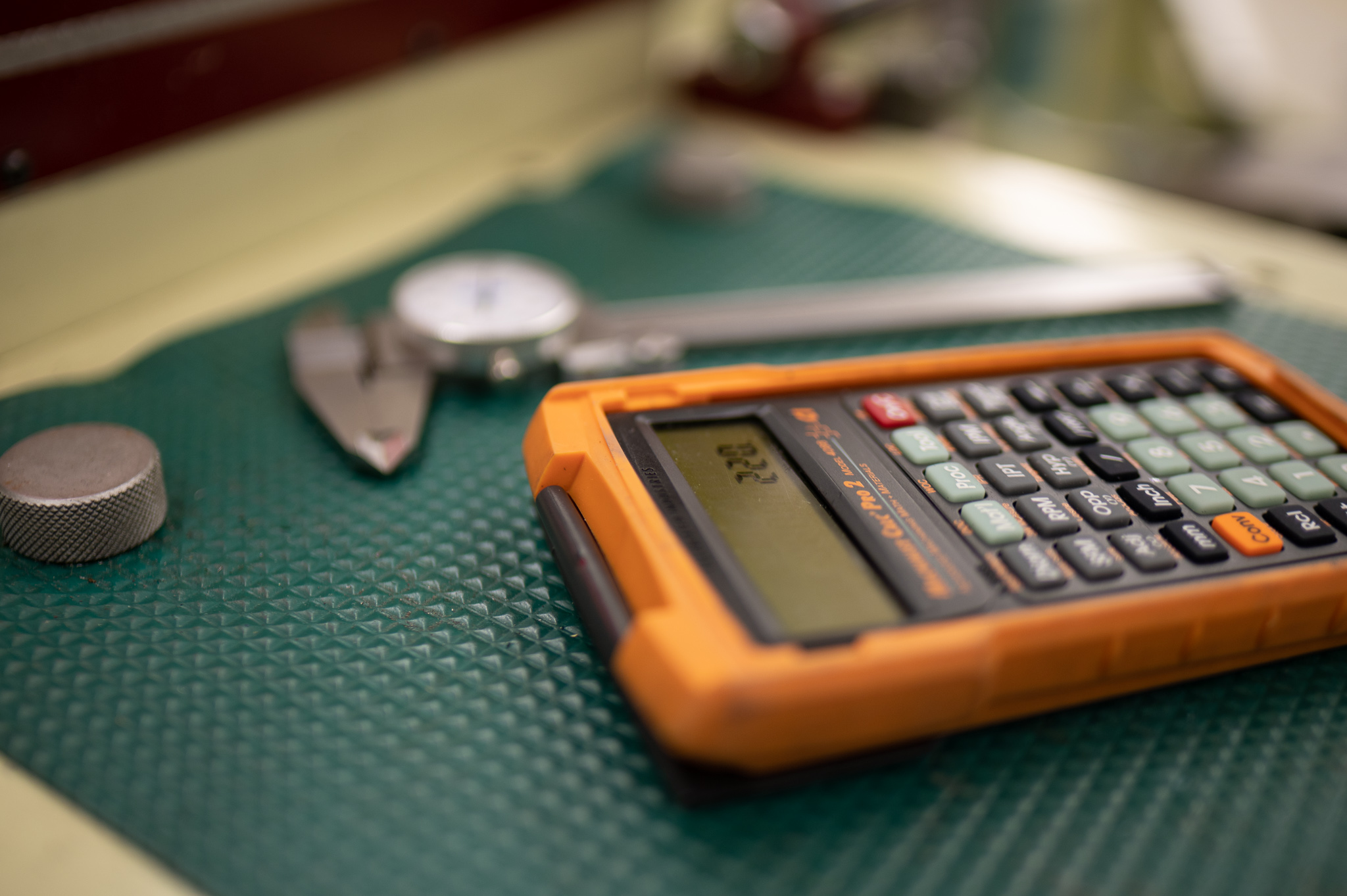 Close-up view of an orange calculator resting on a green textured work surface, with metal measuring tools including a caliper and gauge blurred in the background, highlighting a technical workspace or trades training environment with small precision instruments and tools.