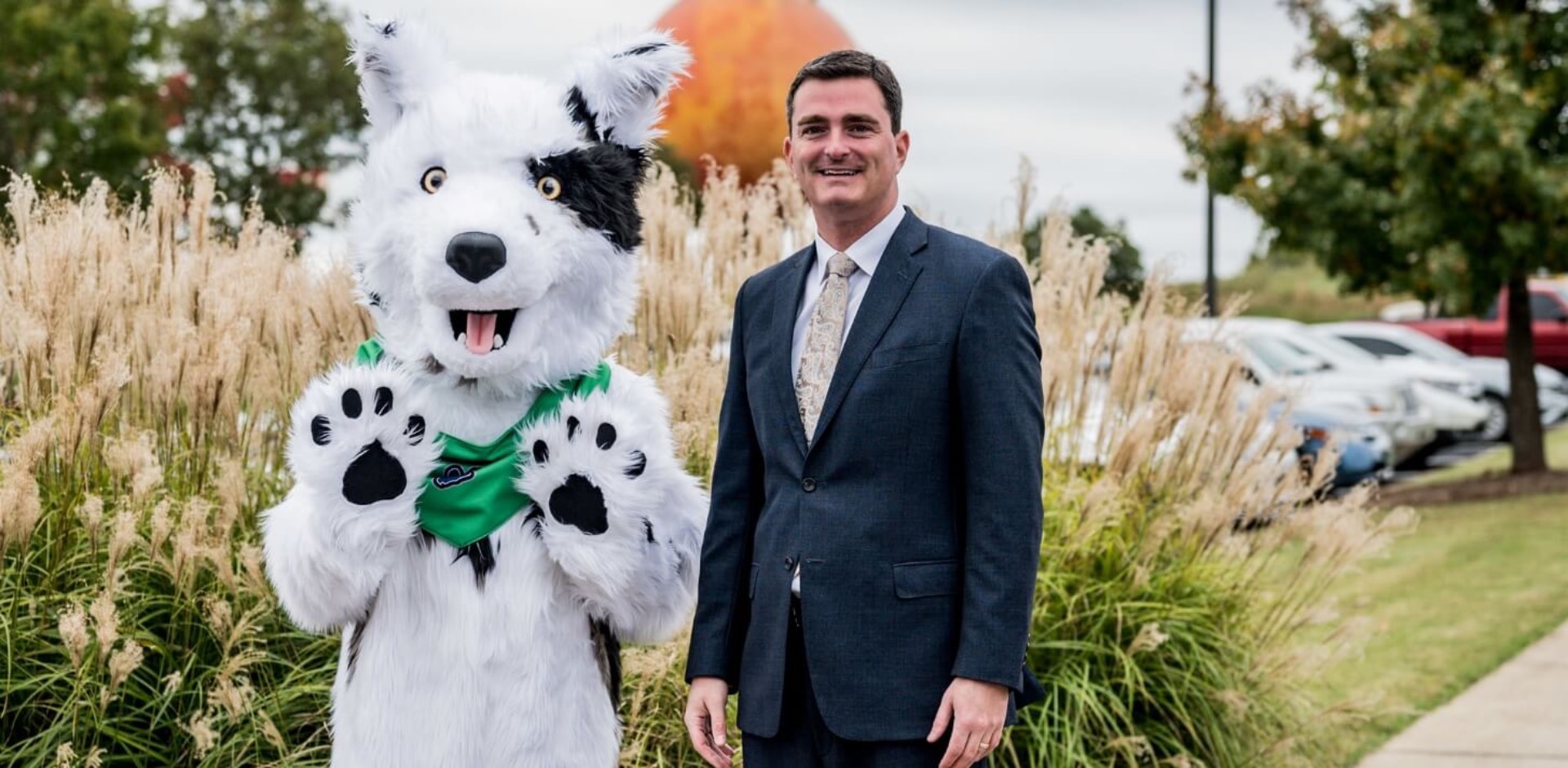 Outdoor campus scene featuring Chaser, the Spartanburg Community College mascot, standing beside an individual in a dark suit along a landscaped walkway with tall grass and parked cars in the background, both facing the camera with raised paws and hands, under soft daylight.
