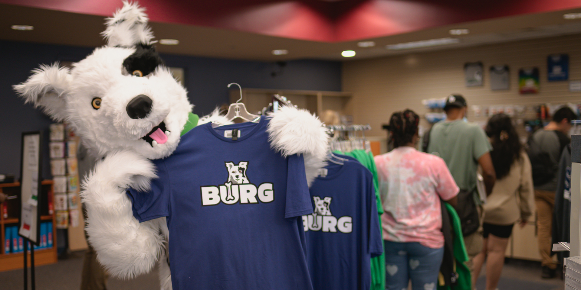 Chaser, the Spartanburg Community College mascot, stands inside a campus bookstore holding a blue “BURG” t-shirt on a hanger, with additional matching shirts displayed on a rack, while several individuals browse merchandise in the background under warm indoor lighting in a retail campus environment.