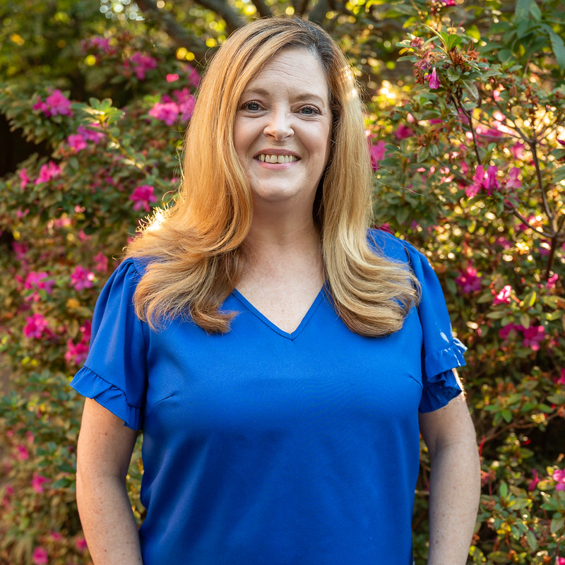 Outdoor portrait of an individual wearing a bright blue blouse with short ruffled sleeves, standing in front of flowering shrubs with pink blossoms and dense green foliage, with natural sunlight creating warm highlights and a softly blurred garden background for a clean professional headshot.