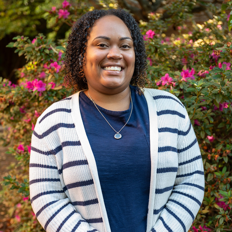 Outdoor portrait of an individual wearing a striped cardigan over a dark shirt, standing in front of flowering shrubs with pink blossoms and green foliage, with natural sunlight creating a softly blurred garden background and a clean, professional campus-style headshot composition.
