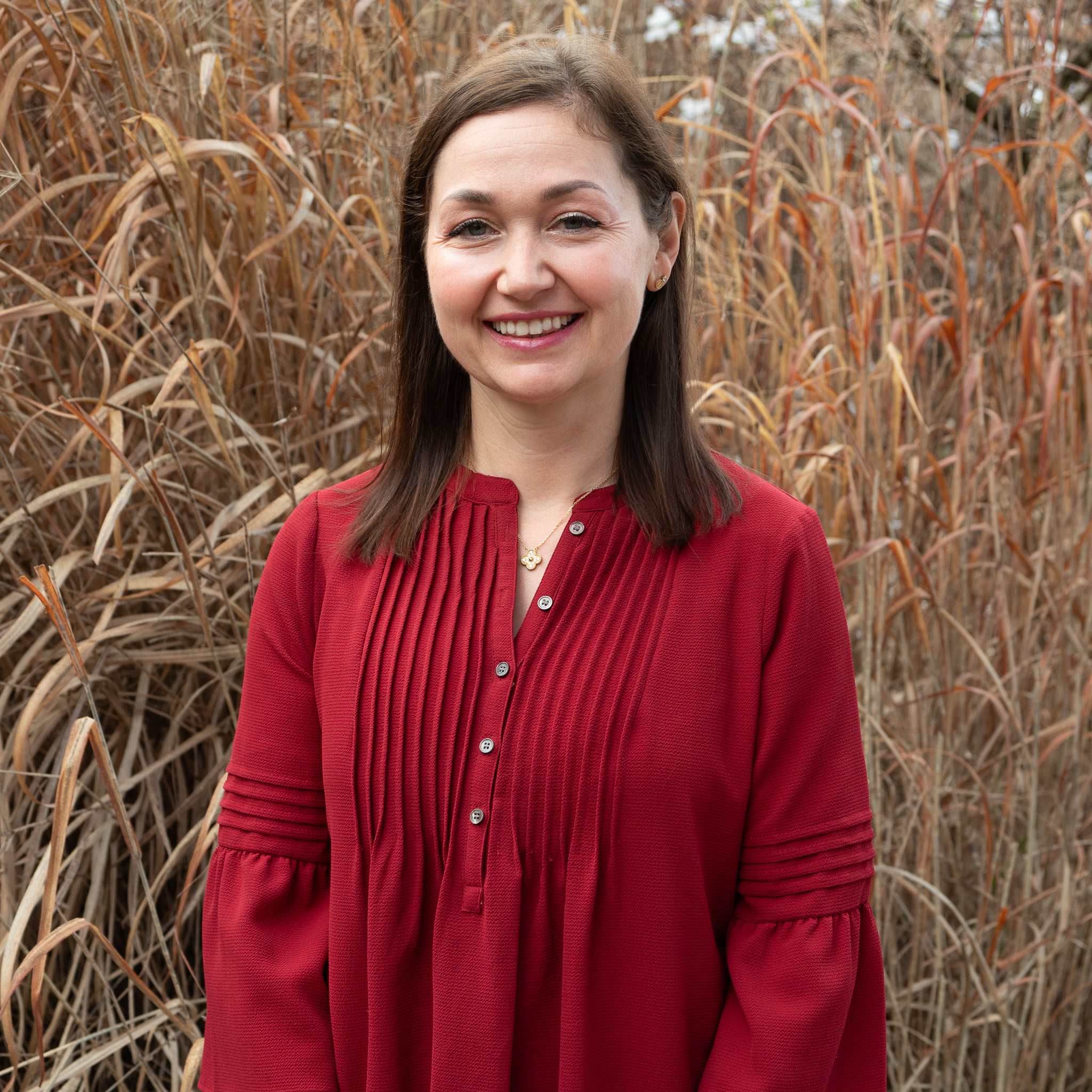 Outdoor portrait of an individual wearing a red textured blouse with pleated front detail and a gold necklace, standing in front of tall dried grasses and natural foliage, with soft daylight illuminating the face and creating a clean, professional campus-style headshot with a neutral, blurred background.
