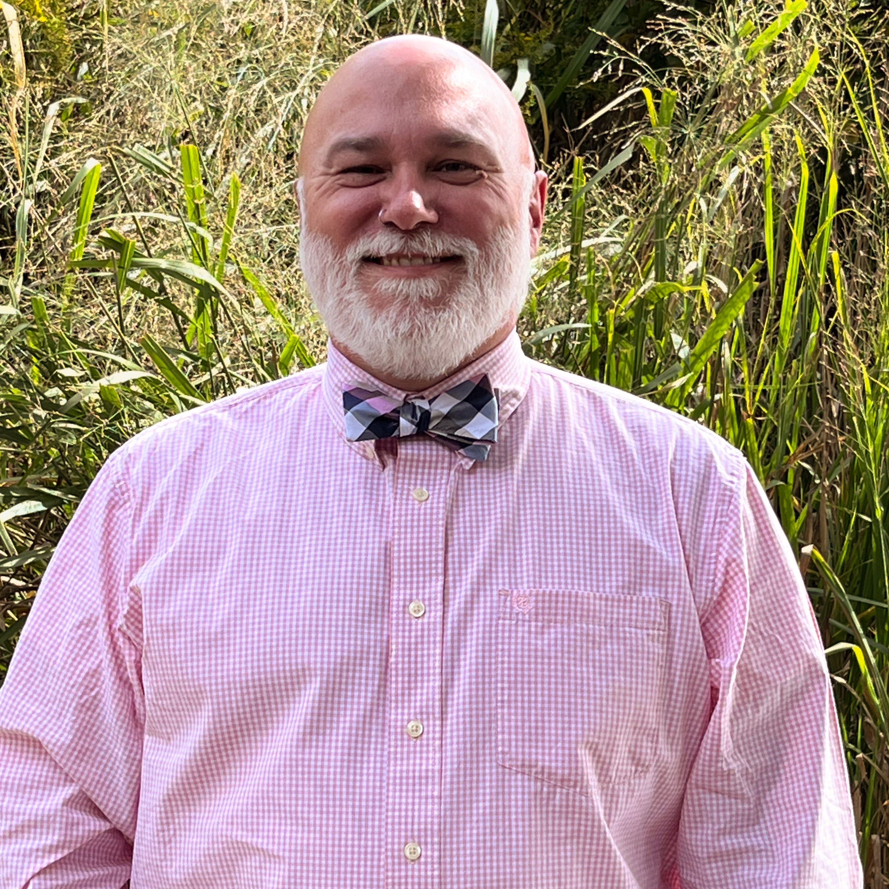 Outdoor portrait of an individual wearing a pink checkered button-up shirt with a patterned bow tie, standing in front of tall green grasses and foliage, with natural sunlight creating bright highlights and a softly blurred background for a clean, professional headshot-style image.